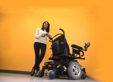 Pooja, founder of BrazeMobility is standing in front of wheelchair smiling.